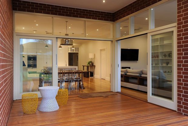 A warm, contemporary open-plan living and dining area opening onto a timber deck. Exposed red brick walls frame extensive internal glazing and sliding doors. The dining area connects to a modern kitchen, while a living room with a sofa and wall-mounted television is visible to the right. Bright woven outdoor stools and a sculptural side table sit on the deck in the foreground.