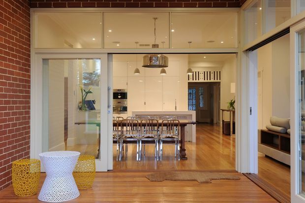 View from a timber-floored enclosed outdoor or transitional area through large glazed sliding doors into a bright open-plan dining and kitchen space. The interior features exposed red-brick walls, warm timber flooring, white built-in kitchen cabinetry, a long dining table with multiple chairs, pendant lighting, and a dark timber doorway beyond. A living area with a sofa is visible through glazing on the right.