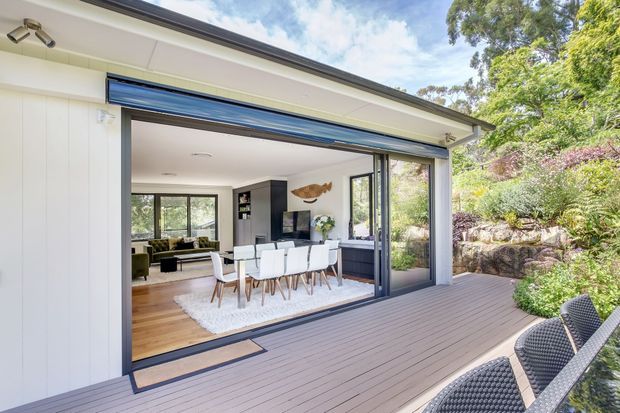 Bright contemporary single-storey home opening onto a spacious timber deck through wide glazed sliding doors. The view shows a dining area and living room inside, with white vertical wall cladding, dark-framed glazing, warm timber flooring, and landscaped rocky garden surrounds.