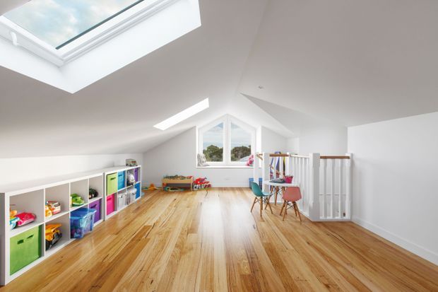 Bright attic-level children's playroom or flexible family room with a sloping white ceiling, large skylights, a central dormer-style window, warm timber flooring, and low open shelving filled with colourful toys and storage bins. A small table and chairs sit near a white stair balustrade, creating an airy, playful space.