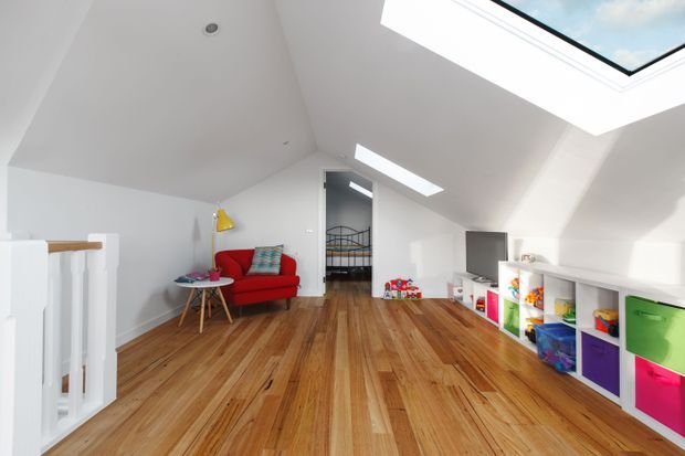 Bright attic-style children's room with a sloped white ceiling, large skylights, warm timber flooring, a red armchair, colourful cushions, open white storage shelving, and a doorway leading to a bedroom. A stair balustrade is visible at the left.