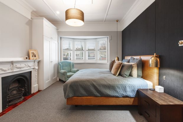 A refined bedroom with a large upholstered bed, layered blue-grey bedding and cushions, a dark textured feature wall, built-in white wardrobes, and a bay window with white plantation shutters. The room also features a decorative white fireplace, grey carpet, timber bedside furniture, brass-toned lighting, and traditional ceiling and wall mouldings.