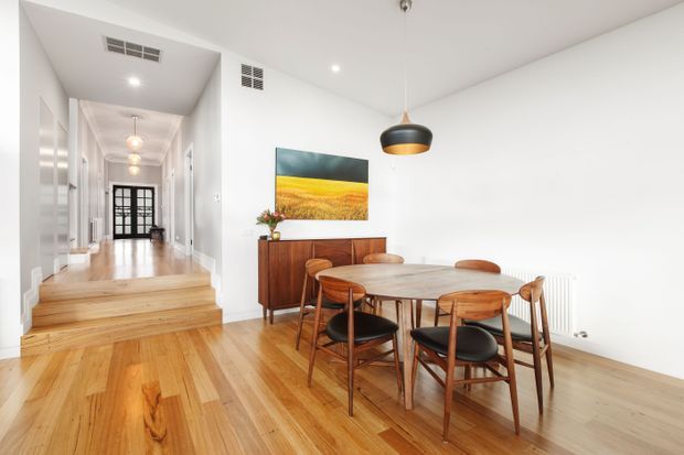 Bright dining area with a mid-century-inspired timber dining table and chairs, a warm timber sideboard, polished timber floors, and a black-and-brass pendant light. The room opens onto a raised hallway through a short set of timber steps, with white walls, high ceilings, and a colourful landscape artwork.