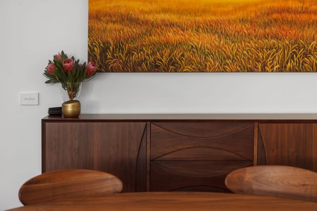 Close-up interior view of a warm timber dining or living area, featuring a dark wood built-in sideboard with sculptural geometric cabinet fronts, a matching timber table and chairs, a small metallic vase with flowers, and a large orange-and-gold abstract artwork on a white wall.