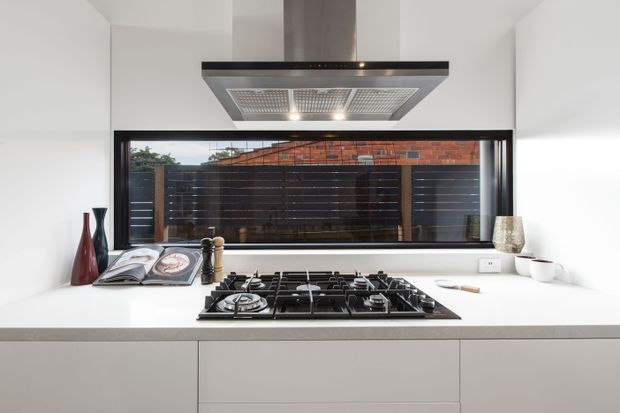 A bright, minimalist kitchen focused on a broad gas cooktop set into a pale benchtop, with a large stainless-steel rangehood above. White flat-panel cabinetry and walls create a clean, streamlined backdrop, while a wide dark-framed window looks onto a timber privacy screen and neighbouring brickwork. Small decorative objects and cookbooks sit along the rear benchtop.