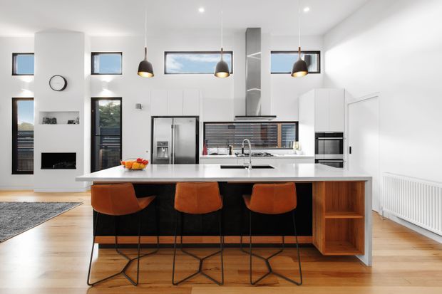 Bright contemporary open-plan kitchen with a large white island, dark cabinetry, integrated appliances, timber flooring, and four burnt-orange bar stools. Pendant lights hang above the island, while a fireplace, glazing, and radiator are visible in the surrounding living area.
