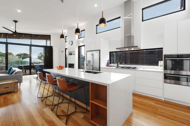Bright open-plan kitchen and living area with a large white island, integrated cabinetry, dark tiled splashback, stainless-steel appliances, timber flooring, pendant lights, and high-level windows. A dining/living zone and glazed outdoor connection are visible beyond the kitchen.