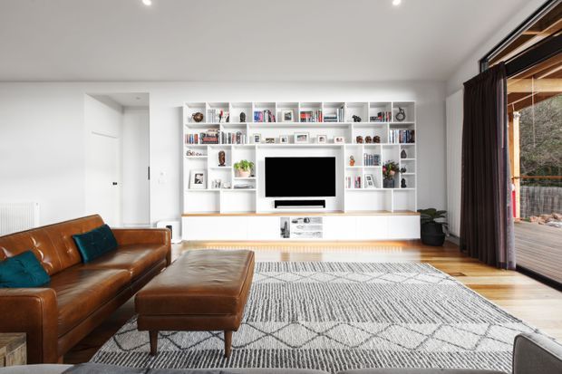 Bright contemporary living room with a large built-in white media and display wall, flat-screen television, extensive open shelving, and concealed storage. A cognac leather sofa and ottoman sit on a large monochrome geometric rug over timber flooring. Full-height glazed doors with dark curtains open to an exterior deck, creating strong indoor-outdoor connection.