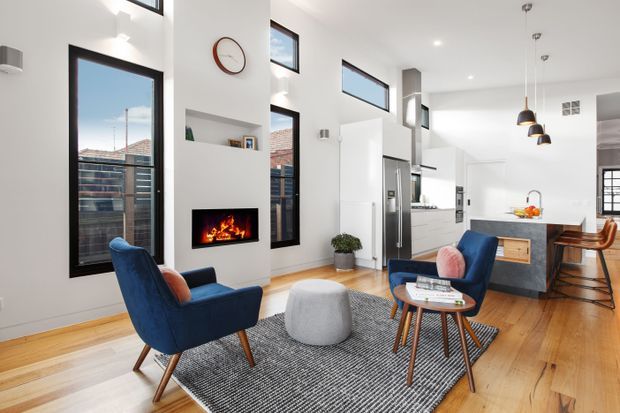 Bright open-plan living room with a double-height white interior, tall dark-framed windows, a built-in fireplace, mid-century-style blue armchairs, a round timber coffee table, and a kitchen with island and dining area beyond. Warm timber flooring and a textured grey rug contrast with the white walls and cabinetry.