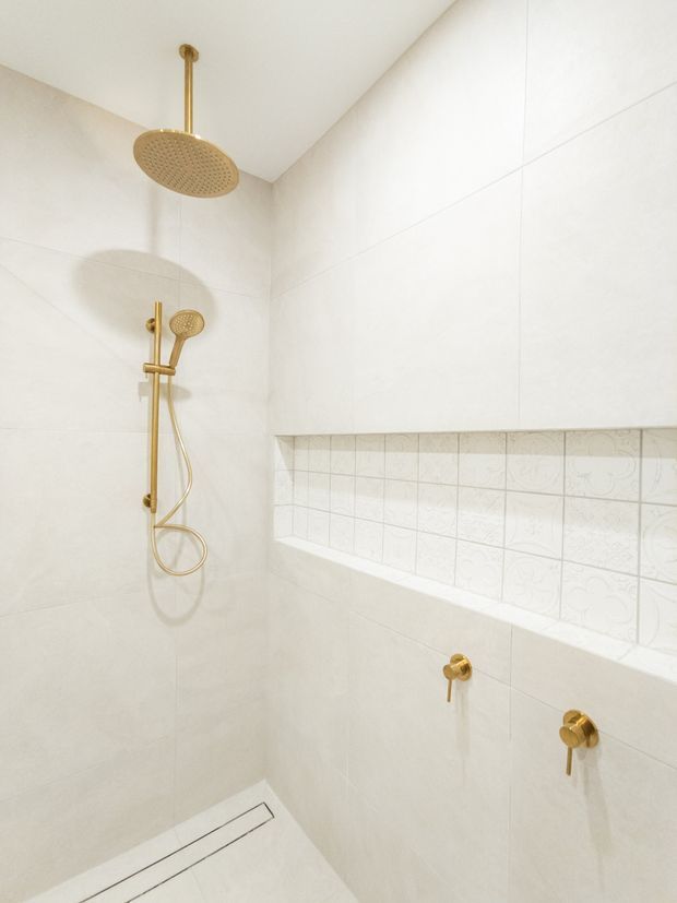A bright, minimalist walk-in shower clad in large off-white tiles, with a recessed niche featuring decorative patterned tiles. Brushed brass/gold rainfall showerhead, handheld shower, controls, and linear floor drain provide warm metallic accents.