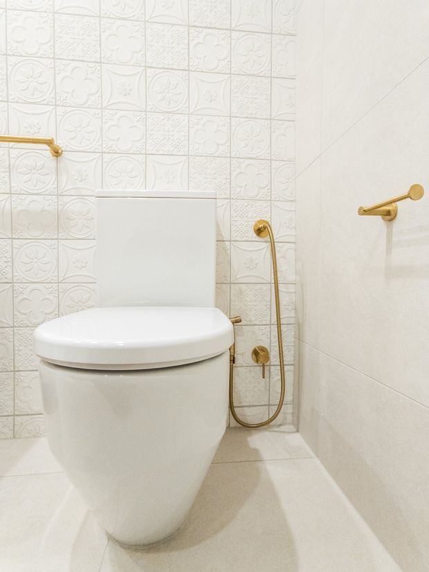 Compact bathroom focused on a white toilet with gold-toned flush controls, bidet spray, and towel rail. The walls and floor are finished in pale tiles; the wall behind the toilet features decorative embossed floral and geometric patterned tiles, while the side wall uses large plain light tiles. The overall scheme is bright, warm, and refined.