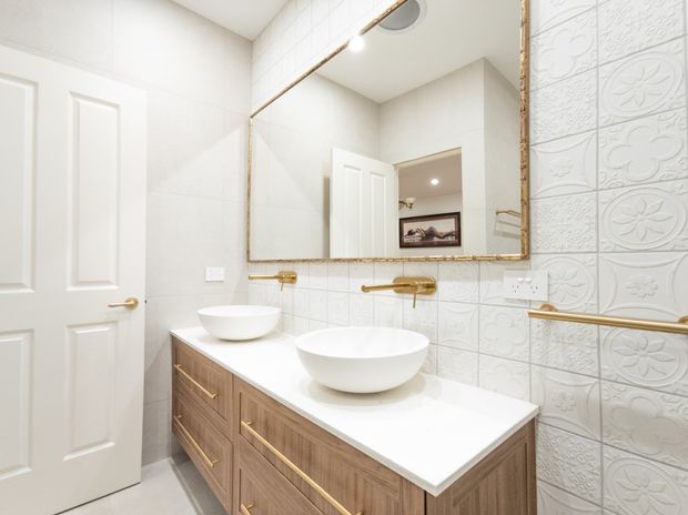 Bright contemporary bathroom with a double-basin timber vanity, white vessel sinks, white benchtop, large framed mirror, white embossed decorative wall tiles, and brass/gold fixtures and hardware. A white paneled door is visible at the left.