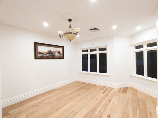 Bright, empty traditional-style room with newly finished light timber flooring, white walls, decorative ceiling cornice and ceiling medallion, a vintage-style chandelier, recessed downlights, and white-framed windows including a projecting bay window. A framed artwork hangs on the wall.