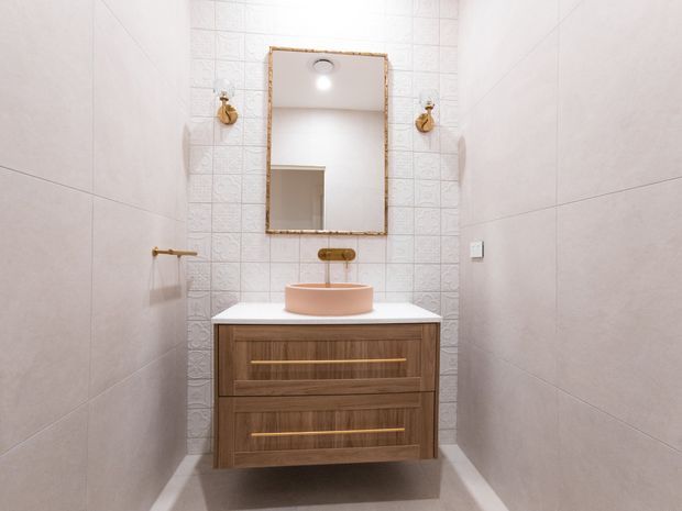 A compact, bright bathroom with large pale wall tiles, a decorative textured tile feature wall, and a floating timber vanity with brass drawer pulls. The vanity has a white benchtop and a round pale pink vessel basin, beneath a framed rectangular mirror with brass-toned detailing. Brass wall sconces and a towel rail add warm metallic accents.