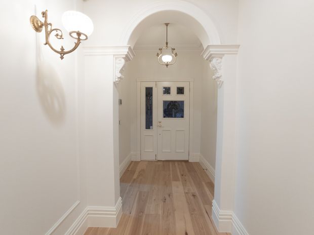 A refined residential entry hall with a white-painted arched opening, ornate decorative capitals and layered traditional cornices, leading to a glazed front door. The space features pale timber flooring, white walls and moulded skirting, with classic brass wall and pendant lights.