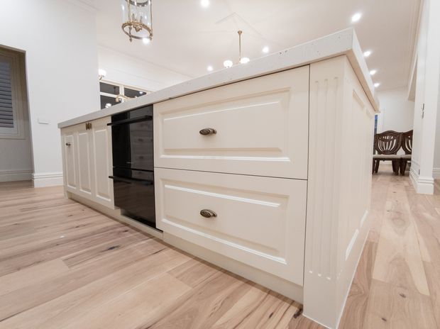 Bright, spacious kitchen centred on a large white painted island with recessed-panel cabinetry, drawers, fluted side detailing, dark integrated appliances, and a pale timber floor. Traditional decorative detailing and pendant lighting are visible, with a dining area beyond.
