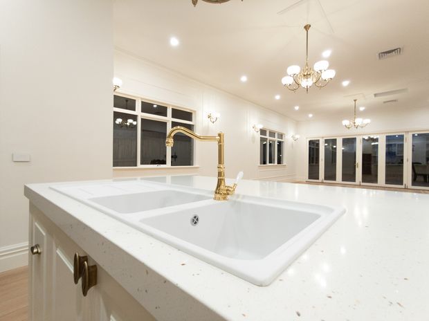 Bright, spacious white kitchen viewed from the sink area, featuring a large white island with an integrated double-basin sink, polished brass gooseneck tap, light cabinetry, extensive windows and glazed doors, recessed lighting, and ornate crystal chandeliers. The space has a refined traditional feel with a clean, light palette.