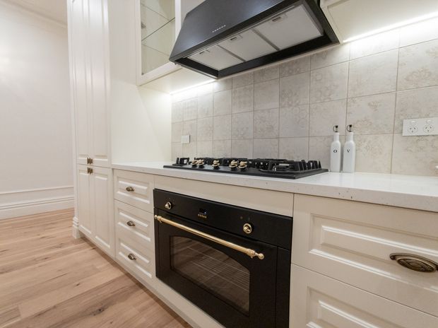 Bright traditional-style kitchen featuring white raised-panel cabinetry, a black freestanding-style oven and gas cooktop with brass hardware, a pale tiled splashback, white benchtop, timber flooring, and a prominent black rangehood.