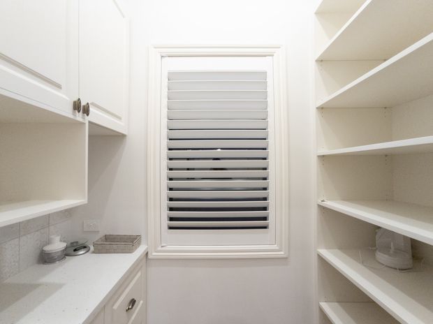 Bright, compact white pantry with built-in cabinetry and open shelving on both sides, a light benchtop, tiled backsplash, and a central window fitted with white plantation shutters. The space has a clean, highly organized, predominantly white finish.