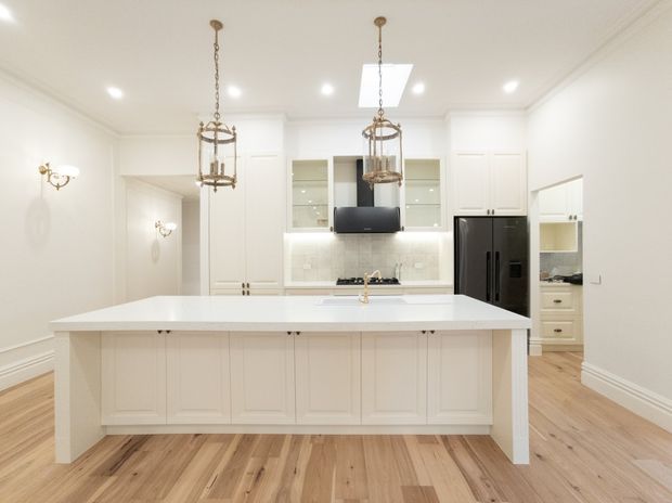 Bright, spacious white kitchen with a large island featuring paneled cabinetry and a white benchtop, integrated sink, shaker-style wall cabinetry, glass-front display cupboards, tiled splashback, black appliances, brass lantern pendant lights, recessed lighting, and light timber flooring. The room has traditional detailing with a refined, clean finish.