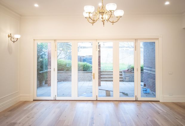 Bright, elegant interior room with polished light timber flooring, cream-painted walls and decorative ceiling cornices. A large set of white-framed glazed folding or French doors opens to a paved courtyard. Ornate brass-and-glass chandeliers and matching wall sconces provide a traditional decorative character.
