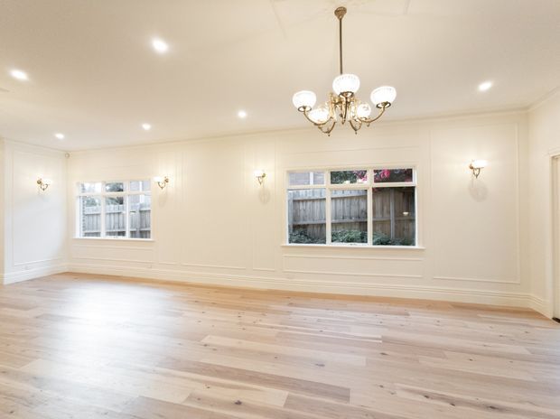 Bright, empty formal interior room with pale wide-plank timber flooring, white paneled walls, decorative cornices and skirting, multiple windows, recessed ceiling lights, wall sconces, and an ornate traditional chandelier. The room appears suitable as a living or dining space, with a refined traditional architectural character.