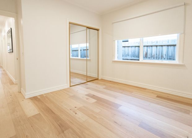 Bright, empty bedroom with pale timber flooring, white walls, mirrored built-in wardrobe doors, a wide window with roller blind, and decorative white skirting and cornices. A hallway is visible through the open doorway.