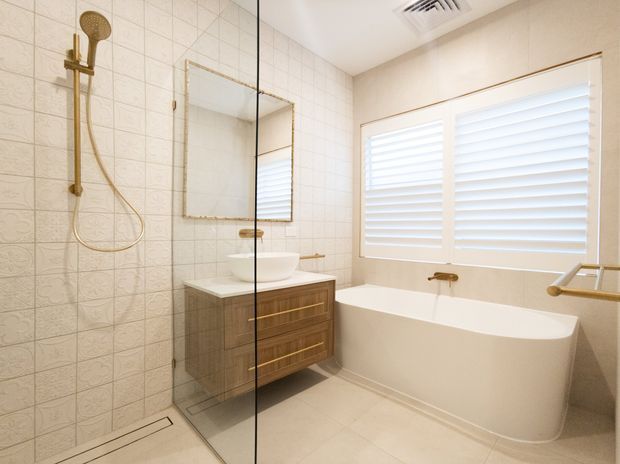 Bright contemporary bathroom featuring a walk-in shower with patterned wall tiles, a floating timber vanity with a vessel basin, a freestanding white bathtub, plantation shutters, frameless glass shower screening, and brushed brass/gold fixtures.