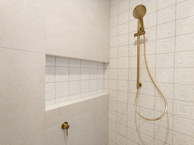 A compact tiled shower area with a wall-mounted brushed gold handheld shower rail and matching control. The walls combine large pale stone-look tiles with small white decorative embossed tiles, and include a recessed tiled niche with a solid ledge.