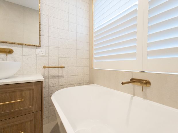 A bright, compact bathroom featuring a freestanding white bathtub beneath a large window with white plantation shutters. The room includes a timber vanity with a white basin and countertop, patterned light wall tiles, brushed brass or gold-toned tapware, and a matching framed mirror.