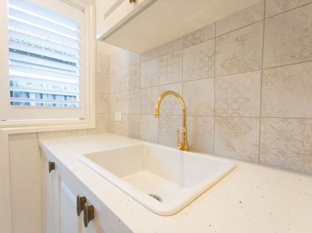 Bright, compact kitchen sink area with a deep white undermount sink set into a white benchtop, white cabinetry, patterned cream-and-beige tiled splashback, and a prominent polished gold gooseneck tap. A white-framed louvred window provides natural light.