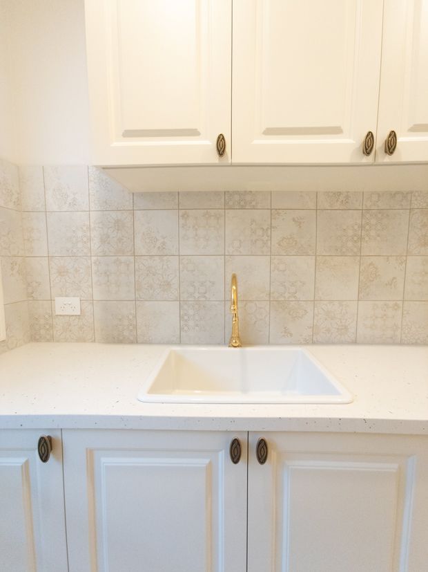 A compact traditional-style kitchen work area featuring white raised-panel cabinetry, ornate dark metal pulls, a white inset sink with a brass-toned faucet, a pale speckled benchtop, and a decorative patterned tiled splashback.