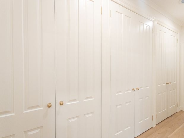 Bright hallway lined with multiple white raised-panel built-in doors and cupboards, featuring small brass knobs, white trim, and pale timber flooring. The corridor has a clean, traditional fitted-joinery appearance.