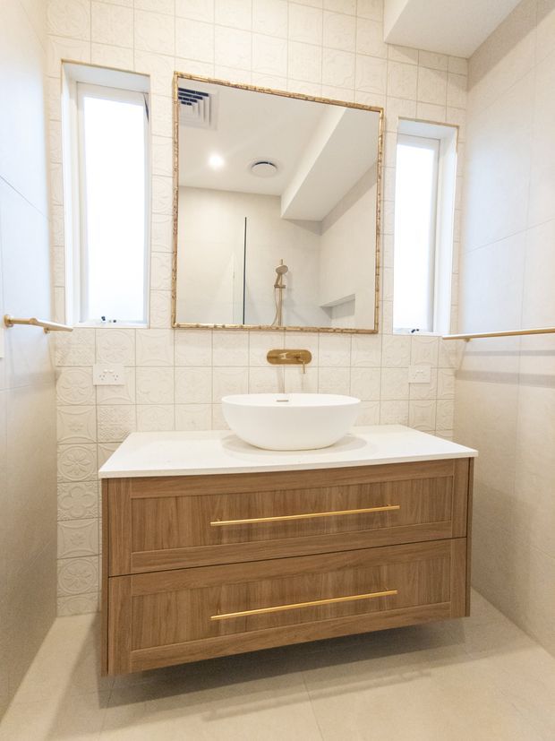 Bright contemporary bathroom featuring a floating timber vanity with a white vessel basin, brass tapware and handles, patterned cream wall tiles, large framed mirror, narrow windows, and a walk-in shower reflected in the mirror.