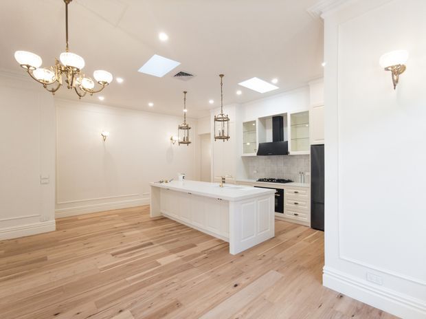 Bright, spacious white kitchen with a large island, shaker-style cabinetry, integrated appliances, marble-look splashback, timber flooring, decorative wall mouldings, chandeliers and pendant lights. The open layout includes a generous dining or living area.