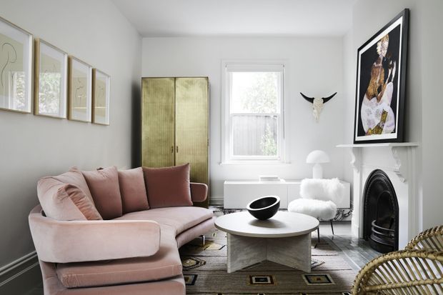 A refined living room with white walls, a soft blush-pink curved sofa, a round light coffee table, patterned area rug, and a black fireplace with a white mantel. The room includes a tall textured cabinet, framed artwork, a sculptural wall-mounted horn display, a white armchair, and a large window providing natural light.