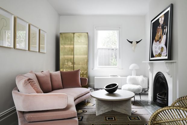 A bright, refined living room with a blush-pink curved sectional sofa, round sculptural coffee table, patterned area rug, white fireplace mantel, black fire surround, framed artwork, decorative wall-mounted horn sculpture, and a glazed cabinet. The room has white walls, timber flooring, layered artwork, and a soft neutral palette with black accents.