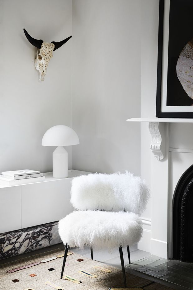 A refined monochromatic living room corner featuring a fluffy white lounge chair, sculptural white dome lamp, low white sideboard, ornate black fireplace, decorative bull skull, framed artwork, patterned stone detail, and a colourful geometric rug. The space combines classic architectural detailing with contemporary sculptural furnishings.