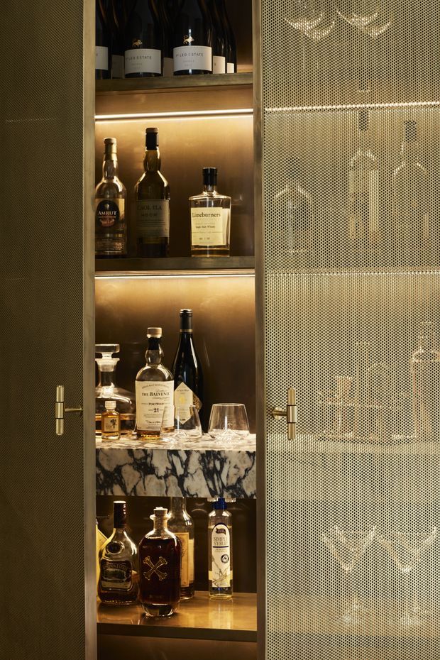 A close-up of a luxurious built-in drinks cabinet displaying wine and spirits on illuminated shelves. The cabinet has dark textured doors with brass handles, warm integrated lighting, and a dramatic black-and-white marble shelf or benchtop detail. One perforated metal door is partially closed, revealing glassware and bottles behind it.