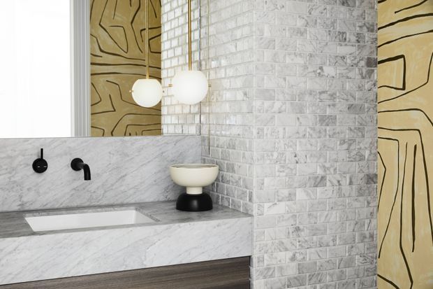 Stylish contemporary bathroom with a sculptural white and black vessel basin, wall-mounted black tapware, integrated marble vanity and backsplash, textured white brick-pattern wall tiles, and warm abstract patterned wall panels. Globe pendant lights add a soft decorative focal point.