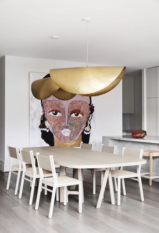 Bright contemporary open-plan dining area with a large pale timber dining table and white dining chairs, dominated by an oversized sculptural gold pendant light. A large colourful portrait artwork forms a focal point behind the table, with a minimalist kitchen and stone island visible in the background.