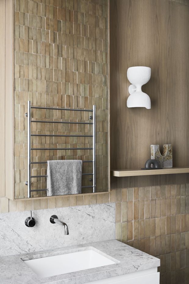 Close-up of a refined contemporary bathroom vanity. The space features a white inset basin set into a pale veined stone benchtop, brushed chrome wall-mounted tapware, warm beige vertically arranged rectangular wall tiles, a chrome heated towel rail, and a dark timber-look recessed shelving wall with a sculptural white wall light and decor.