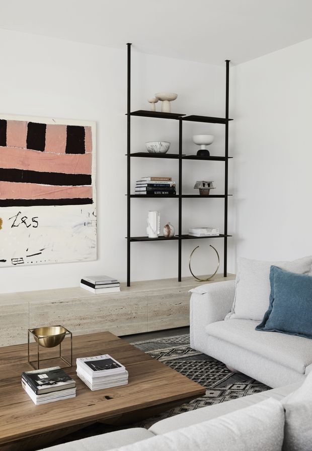 A bright, refined living room with a white plastered interior, a slim black open shelving unit displaying ceramics and books, a pale stone or concrete built-in media cabinet, abstract wall artwork, and soft white sofas. A warm timber coffee table, patterned rug, blue cushion, and brass-toned side table add contrast and texture.