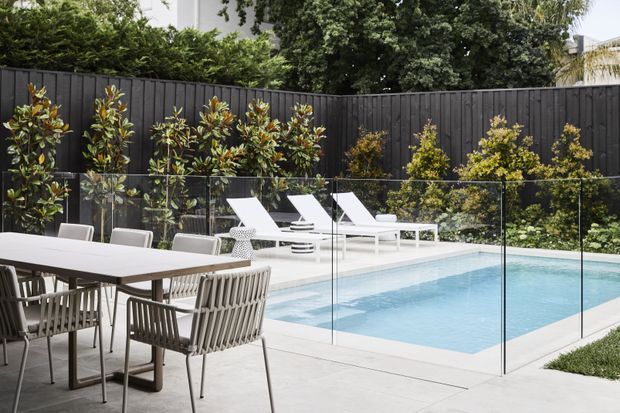 Contemporary outdoor entertaining area beside a rectangular swimming pool, with a large dining table and woven chairs in the foreground, white sun loungers beyond the frameless glass pool fence, and layered planting against a dark vertical timber fence.