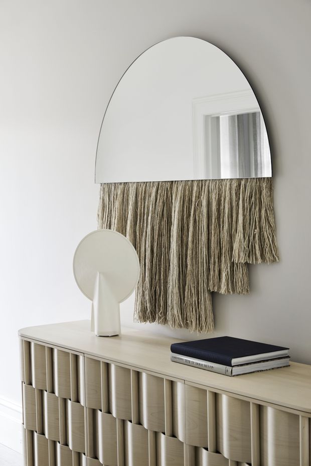 A refined neutral-toned interior vignette featuring a half-round wall mirror with layered hanging textile fringe beneath it, mounted above a light timber fluted sideboard. A small sculptural white lamp and stacked books sit on the sideboard. The composition is softly lit and textural, with a calm, contemporary decorative style.