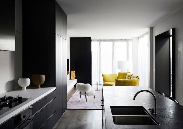 A sleek open-plan contemporary interior combining a dark, minimalist kitchen with a bright living area. The kitchen features matte black cabinetry, a dark stone island with integrated double sinks and a curved black tap, light timber flooring, and a pale stone or composite benchtop. Beyond the kitchen is a refined living space with floor-to-ceiling sheer glazing, a pale rug, sculptural seating, and mustard-yellow sofas.