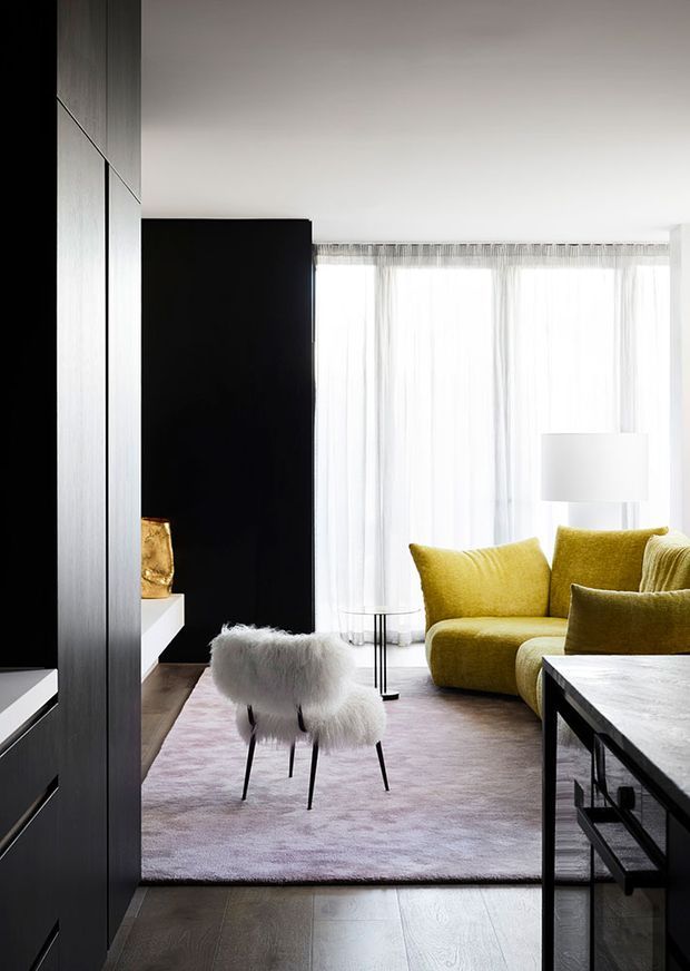 A bright, contemporary living room with floor-to-ceiling sheer curtains, a mustard-yellow sofa, a white floor lamp, a pale textured rug, and a small furry upholstered stool. Dark vertical built-in cabinetry and a black partition frame the room, while a dark stone or composite coffee table occupies the foreground. The space has a refined, minimalist feel with strong black-and-white contrast softened by warm yellow upholstery and timber flooring.