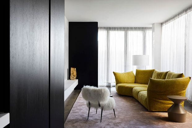 A refined contemporary living room with a mustard-yellow sectional sofa, sculptural side table, white sheepskin accent chairs, and a large pale rug. Floor-to-ceiling sheer curtains wrap around the bright corner glazing, while dark timber or black wall panelling and a black fireplace or media wall create strong contrast.