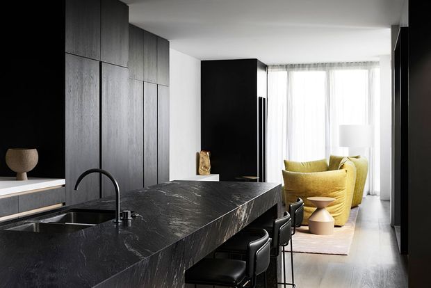 A refined open-plan kitchen with a large dark stone island and integrated sink, black cabinetry and tall timber-grain joinery. Three black bar stools line the island, while a bright living area with mustard-yellow lounge chairs is visible beyond, framed by sheer floor-to-ceiling curtains. The palette is predominantly black, charcoal, timber and warm yellow, creating a sophisticated contemporary interior.