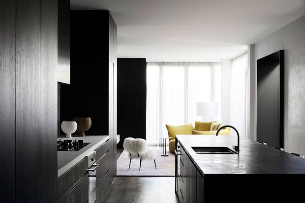 A sleek open-plan kitchen opening into a bright living area. The kitchen features dark timber cabinetry, a large dark stone island with an integrated sink and black tapware, and a pale timber or stone floor. Beyond the kitchen are a neutral sofa, sculptural chairs, and floor-to-ceiling sheer curtains, creating a high-contrast, refined contemporary interior.